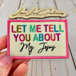 Può includere: Una toppa rettangolare con la scritta ricamata "LET ME TELL YOU ABOUT My Jesus" in vari colori. La toppa ha uno sfondo color crema con un bordo rosa.