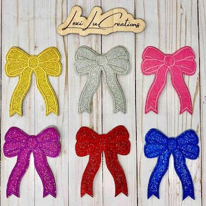 Może przedstawiać: Sześć brokatowych aplikacji w kształcie kokardek w kolorach złotym, srebrnym, różowym, fioletowym, czerwonym i niebieskim. Kokardy ułożone są na jasnej powierzchni z usłojeniem drewna. U góry znajduje się drewniany znak z napisem "Lexi Lu Creations".