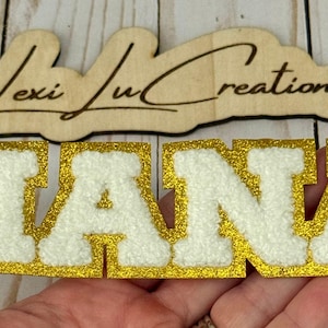 May include: A gold glitter-edged patch spelling "NANA" in white, textured letters. Above it, a wooden sign with the text "Lexi Ju Creations" in a cursive font. The patch is likely for crafting or personalization.