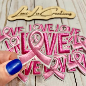 May include: Embroidered pink ribbon patches with the word "LOVE" in a bold font. The patches are on a white background with a light pink border. A wooden sign with the text "Lexi Ju Creations" is in the background.