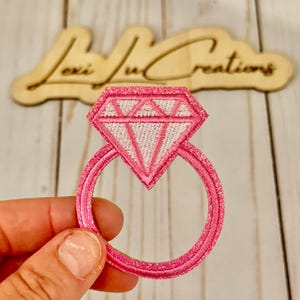 Puede incluir: Un parche bordado rosa con forma de anillo de compromiso de diamantes. El anillo presenta un contorno rosa brillante y un diamante bordado blanco. El parche es probablemente para ropa o accesorios.