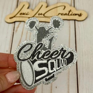 May include: A glittery patch with the words "Cheer Squad" in black script and block letters, with a cheerleader graphic. The patch has a silver glitter border and is held by a hand.