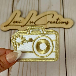 May include: White and gold embroidered camera patch. The patch has a gold outline and detailing, including the lens, flash, and camera body. The text "lexi lu creations" is visible in the background.
