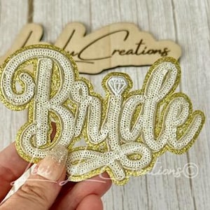 May include: A decorative patch featuring the word Bride in white sequins with a gold sequin border and a small diamond ring icon above the letter i. The patch has a script font style and is designed with a layered appearance, showcasing a gold glitter backing.