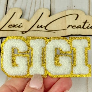 May include: A white and gold patch with the name "GIGI" in a varsity letter style. The patch has a gold glitter border and a fluffy white center. The wooden sign above reads "Lexi Lu Creations".
