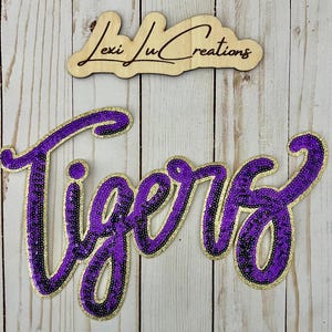 May include: Purple sequined "Tigers" word art with a gold glitter outline. A wooden sign with the text "Lexi Lu Creations" is above the word art. The background is a light wood grain.