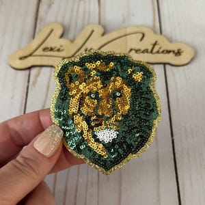 May include: A sequined lion head patch in green and gold. The lion's mane is green, and the face is gold. The patch is outlined in gold glitter. The background is a light wood surface.