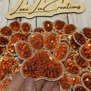 May include: A pile of orange and silver sequined paw print iron-on patches.  The patches are approximately 2 inches in diameter.