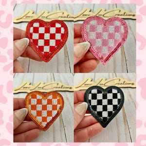Op de afbeelding: Vier hartvormige patches met een dambordpatroon. De patches zijn rood, roze, oranje en zwart, elk met een dambordontwerp in een andere kleur. De randen zijn omrand met pailletten. De patches worden door een hand omhoog gehouden.