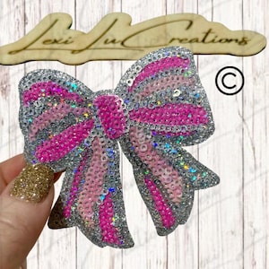 May include: A sparkly bow with pink and silver sequins. The bow has a layered design with alternating pink and silver stripes. The bow is held by a hand with glittery gold nail polish. The wooden sign above the bow says "Lexi Lu Creations".