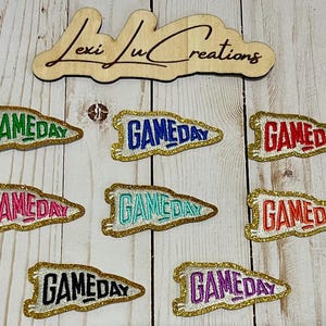 May include: Eight iron-on patches with gold glitter trim and the word "GAMEDAY" in different colors, including red, blue, green, pink, black, and purple.