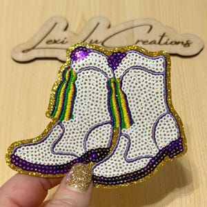 Puede incluir: Aplique en forma de bota blanca con lentejuelas doradas, con detalles en morado, verde y amarillo. El aplique está adornado con pequeños brillantes. El recorte de madera en el fondo dice "Lexi Lu Creations."