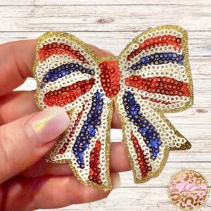 May include: A decorative bow applique covered in shimmering sequins arranged in alternating stripes of red, white, and blue. The edges of the bow are outlined in gold glitter, and the center knot is composed of solid red sequins.