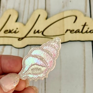 Op de afbeelding: Een geborduurde schelppatch met iriserende en roze tinten. De patch heeft de vorm van een zeeschelp, met gedetailleerde stiksels. De achtergrond toont een houten bord met de tekst "Lexi Lu Creations".