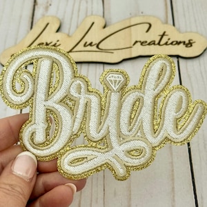 May include: Embroidered patch with the word "Bride" in elegant script, featuring a diamond accent. The patch has a white embroidered design with a gold glitter border. A wooden sign with the text "Lexi Lu Creations" is in the background.