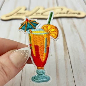 Pode incluir: Patch bordado de um coquetel em um copo alto. A bebida é um gradiente de vermelho, laranja e amarelo, com um canudo verde, uma fatia de laranja e um pequeno guarda-chuva azul. O patch tem uma borda branca.