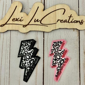 May include: Two embroidered lightning bolt patches. One is black with a silver leopard print, and the other is pink with a leopard print and red sequins. The wooden sign above reads "Lexi Lu Creations".