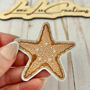 Puede incluir: Parche bordado de estrella de mar en tonos beige y marrón, con borde blanco. El parche se sostiene en una mano. El fondo incluye un letrero de madera con el texto "Lexi Lu Creations".