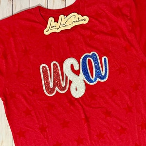 May include: Red t-shirt with a white star pattern and a glittery red, white, and blue "USA" applique.