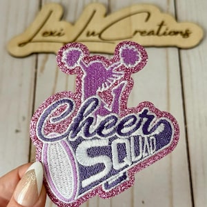 May include: A glittery purple and pink embroidered patch with the words "Cheer Squad" in a stylized font. The patch features a cheerleader graphic and is likely for apparel or accessories.