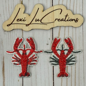 May include: Two red sequined lobster patches with silver glitter trim. The patches are on a light wood grain surface. Above the patches is a wooden sign with the text "Lexi Lu Creations" in a cursive font.