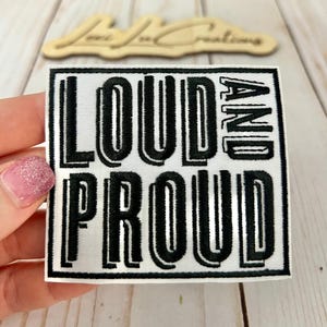 Puede incluir: Parche cuadrado blanco bordado con las palabras "LOUD AND PROUD" en letras mayúsculas negras y en negrita. El parche tiene un borde negro. El parche lo sostiene una persona con esmalte de uñas rosa.