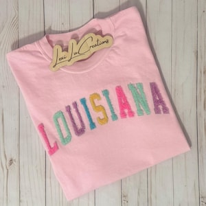 Può includere: Una felpa rosa chiaro con la scritta "LOUISIANA" ricamata in lettere colorate e glitterate. La felpa ha un girocollo ed è piegata su una superficie di legno chiaro. Un'etichetta di legno con la scritta "Lexi Lu Creations" è sul colletto.