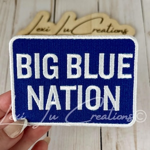 May include: A rectangular embroidered patch with a royal blue background and a white border. The text BIG BLUE NATION is stitched in bold white capital letters across the center of the patch. The patch has a textured fabric surface and clean edges.
