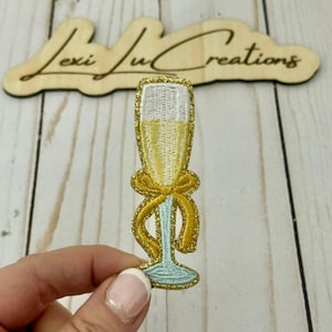 Peut inclure: Écusson brodé représentant une flûte à champagne avec des paillettes dorées, un nœud jaune et une base et un pied bleu clair. Le verre est rempli d'un liquide jaune. Le texte "Lexi Ju Creations" est en arrière-plan.