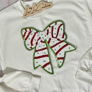 Pu&ograve; includere: Felpa bianca con un grande design a fiocco con paillettes. Il fiocco presenta strisce rosse e un contorno verde, su sfondo bianco. La felpa ha maniche lunghe e girocollo. Un cartello di legno con la scritta "Lexi Lu Creations" &egrave; in alto.