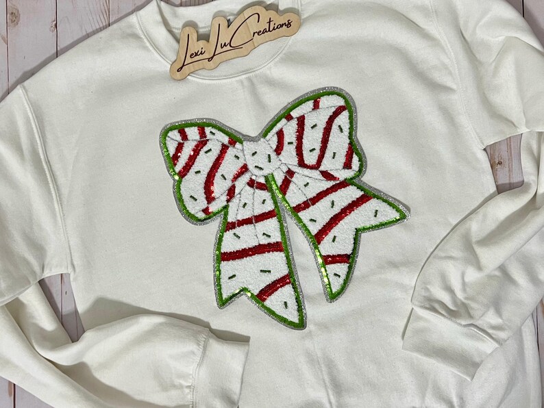 Pu&ograve; includere: Felpa bianca con un grande design a fiocco con paillettes. Il fiocco presenta strisce rosse e un contorno verde, su sfondo bianco. La felpa ha maniche lunghe e girocollo. Un cartello di legno con la scritta "Lexi Lu Creations" &egrave; in alto.