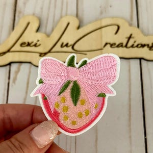 May include: Embroidered strawberry patch with a pink bow, green leaves, and yellow seeds. The patch is on a white background. The wooden sign behind the patch says "Lexi Ju Creations".