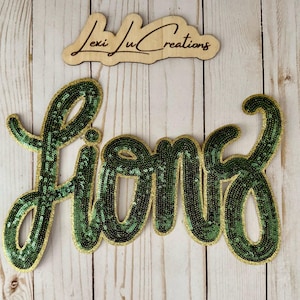 May include: A green sequined "Fiona" word art piece with a gold border, displayed on a light wood background. Above it is a wooden sign that reads "Lexi Ju Creations" in cursive.