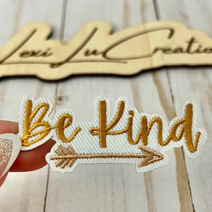 May include: Embroidered patch with the words "Be Kind" in gold thread, with an arrow design. The patch is on a white background. A wooden sign with the text "Lexi Lu Creations" is in the background.