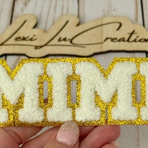 May include: A gold glitter patch spelling "MIMI" in white, fluffy letters. The patch is rectangular and is held by a person. A wooden sign with the text "Lexi Lu Creations" is in the background.