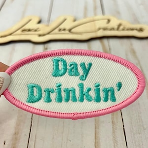 Op de afbeelding: Ovaal geborduurde patch met de woorden "Day Drinkin'" in turquoise letters. De patch heeft een witte achtergrond en een roze gestikte rand. De patch wordt door een hand vastgehouden, met een houten bord op de achtergrond.