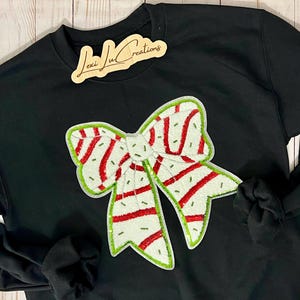 Pu&ograve; includere: Felpa nera con un grande design a fiocco ricamato. Il fiocco presenta una base bianca con strisce rosse e bordi verdi. Il testo "Lexi Ju Creations" &egrave; su un'etichetta di legno.