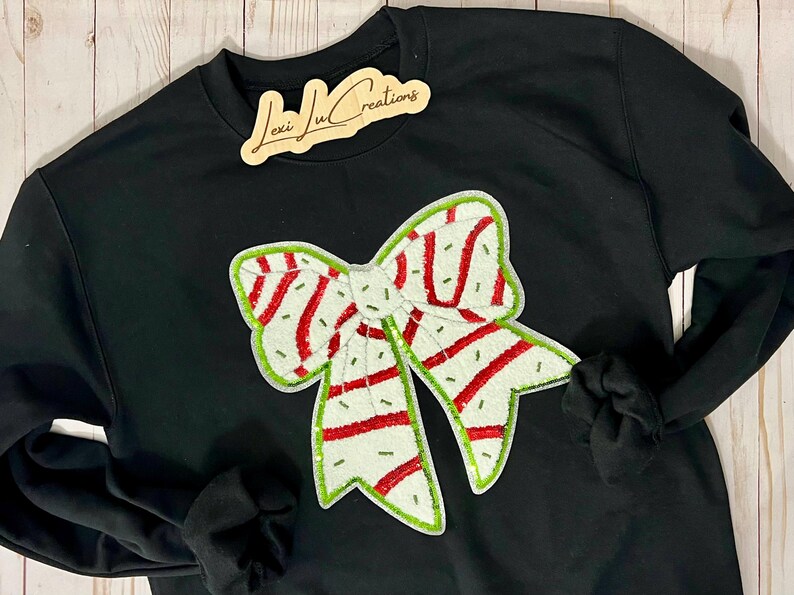 Pu&ograve; includere: Felpa nera con un grande design a fiocco ricamato. Il fiocco presenta una base bianca con strisce rosse e bordi verdi. Il testo "Lexi Ju Creations" &egrave; su un'etichetta di legno.