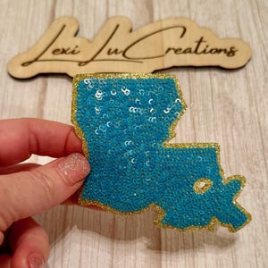 May include: A sequined patch in the shape of Louisiana, filled with turquoise sequins and outlined in gold glitter. The patch is held up in front of a wooden sign that says "Lexi Ju Creations".