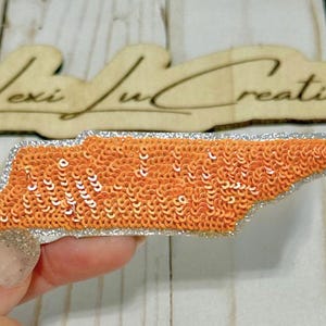May include: An orange sequined patch in the shape of the state of Tennessee with a silver glitter border. The patch is held in a hand. A wooden sign with the text "Lexi Lu Creations" is in the background.