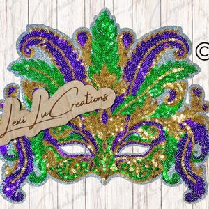 May include: A decorative Mardi Gras mask with a glittery silver border. The mask is adorned with sequins in purple, green, and gold. A wooden sign with the text "Lexi Lu Creations" is placed on the mask.