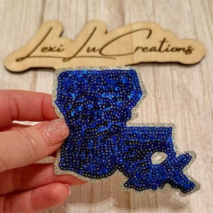 May include: A blue sequined Louisiana state-shaped patch with a silver glitter border. The patch is held in a hand, with a wooden sign that reads "Lexi Lu Creations" in the background.
