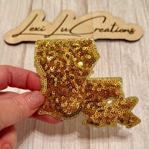 May include: A gold sequined Louisiana state-shaped patch. The patch is filled with small, shiny gold sequins and has a gold border. The background features a wooden sign with the text "Lexi Ju Creations".