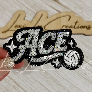 May include: A black patch with white sequin lettering that spells out the word ACE. The design features a white sequin volleyball icon and small white sequin star accents. The patch has a glittery black background and a thick black border.