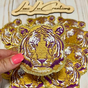 May include: A set of gold and purple embroidered iron-on patches featuring a tiger face design.