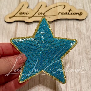 May include: A turquoise sequined star-shaped patch with a gold glitter border. The patch is held by a hand. The wooden sign in the background reads "Lexi Lu Creations".