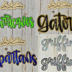 May include: Wooden cutouts featuring the names "Hurricanes", "Gators", "Spartans", and "Griffins" in glittery script. Each name is a different colour, including green, black, blue, and silver. The "Lexi Ju Creations" logo is above each name.