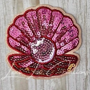 May include: A sequined seashell patch in shades of pink and red. The shell is open, revealing a pearl-like center. The edges are outlined in a darker red, with a gold border. The patch is likely for embellishing clothing or accessories.