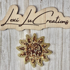 May include: A wooden sign with the text "Lexi Lu Creations" in a cursive font. Below the sign is a gold and brown sequined sunflower applique.