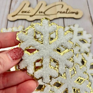 May include: White snowflake ornament with gold glitter trim. The snowflake has a textured, fuzzy appearance. The background includes other snowflakes and a wooden sign that reads "Lexi Jul Creations".
